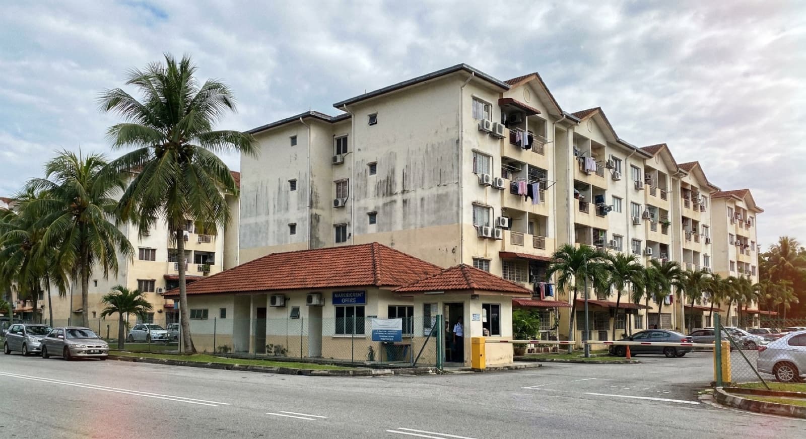 Mid-tier Puchong residensi — weathered off-white paint, air-con units, laundry drying on balconies, guardhouse at entrance