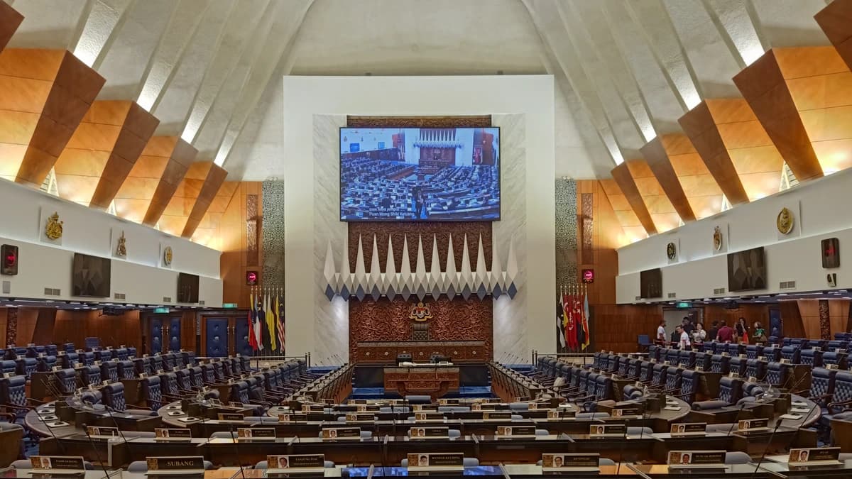 The Dewan Rakyat chamber in Malaysia's Parliament with rows of desks, name plates, flags and a large screen under an angular timber ceiling