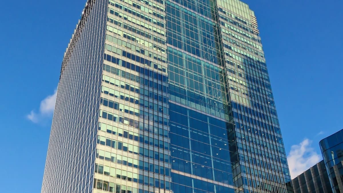 A tall glass office tower with a grid facade rises against a blue sky above Canary Wharf