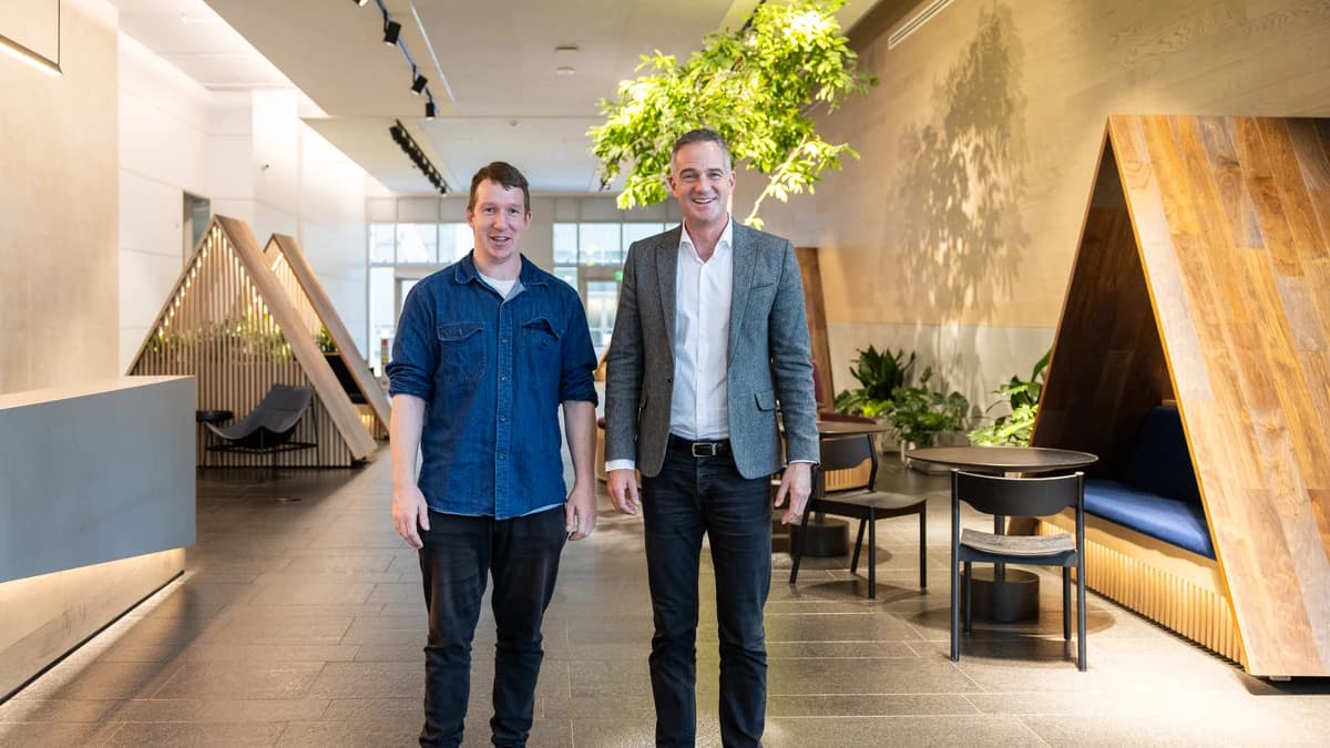 Two men stand side by side in a bright wood-panelled lobby with a hanging plant and seating booths behind them