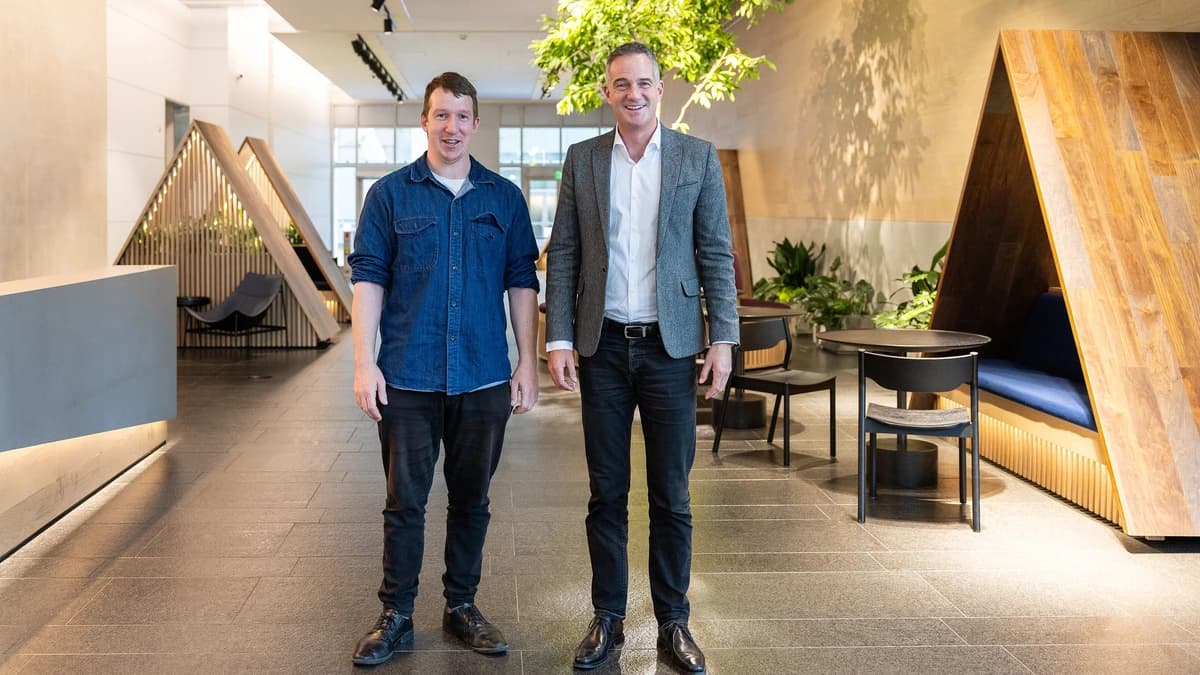 Two men stand side by side in a bright office lobby with timber A-frame seating pods, a dark stone floor and a small tree under track lighting