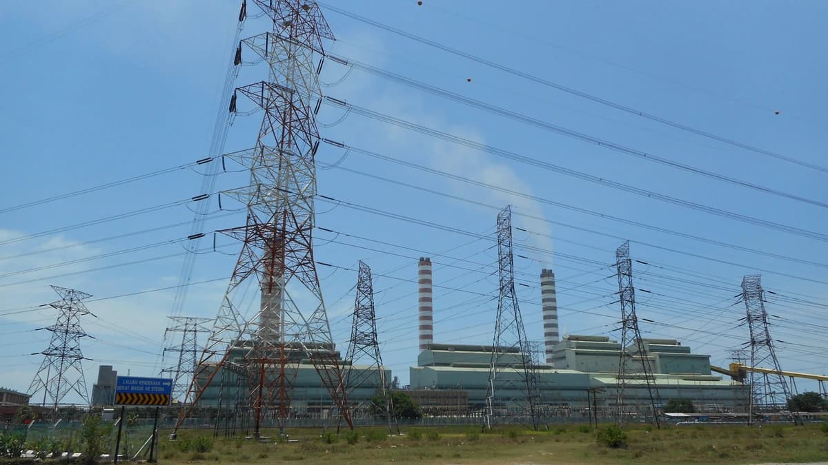 Steel transmission towers and power lines in front of a large coal-fired power station with two striped chimneys under a blue sky