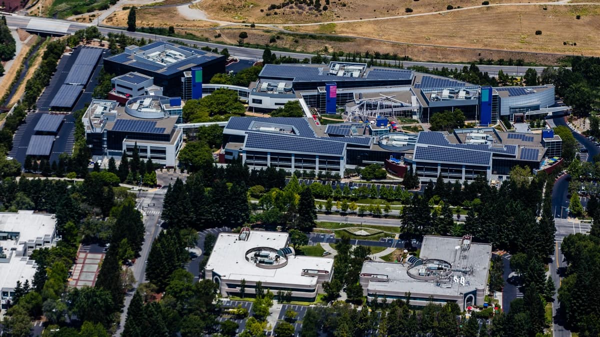 Aerial view of the Google campus in Mountain View, California, with low office buildings, solar panels on the roofs, tree-lined paths and dry hills behind