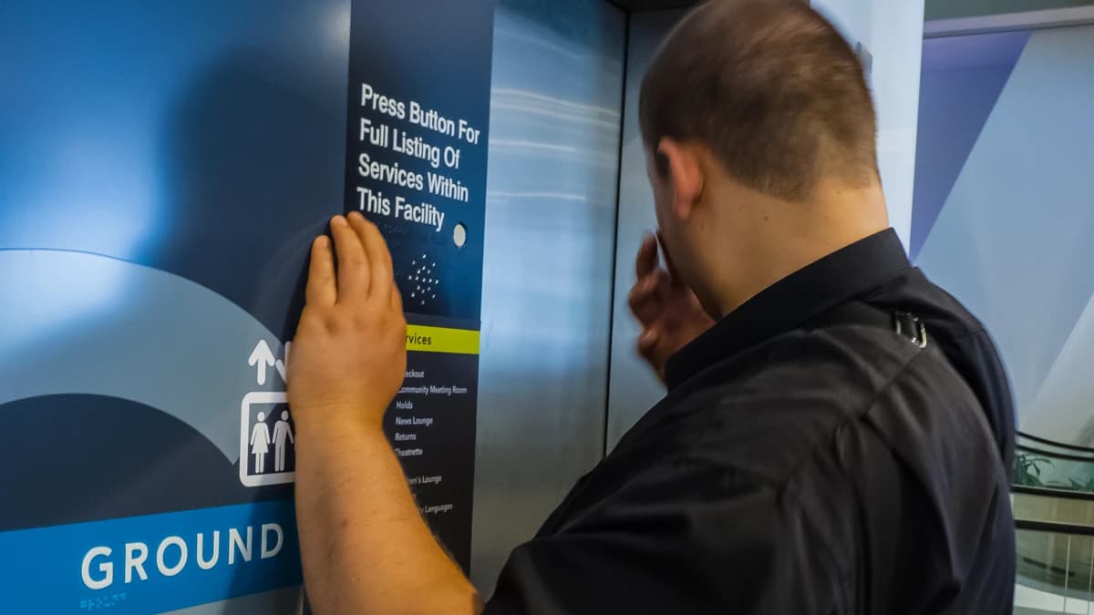 A man in a black shirt rests one hand on a braille and print directory sign beside a lift, with a white cane visible at the lower right