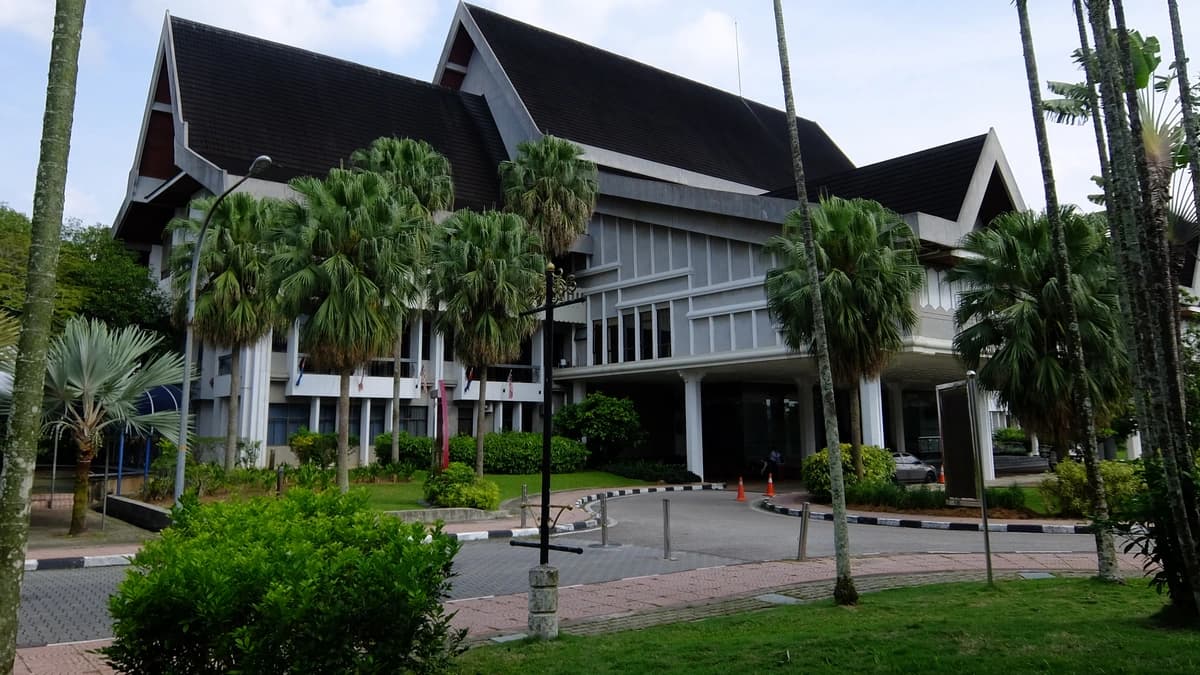 The Sultan Ibrahim Chancellor Building at UTM Skudai, a white building with a dark pitched roof and wide eaves, framed by tall palm trees and a paved path