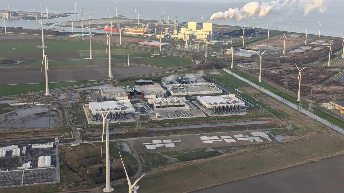 Aerial view of several white Google data centre halls in flat Dutch farmland, ringed by wind turbines, with a power station releasing steam near the coast behind them