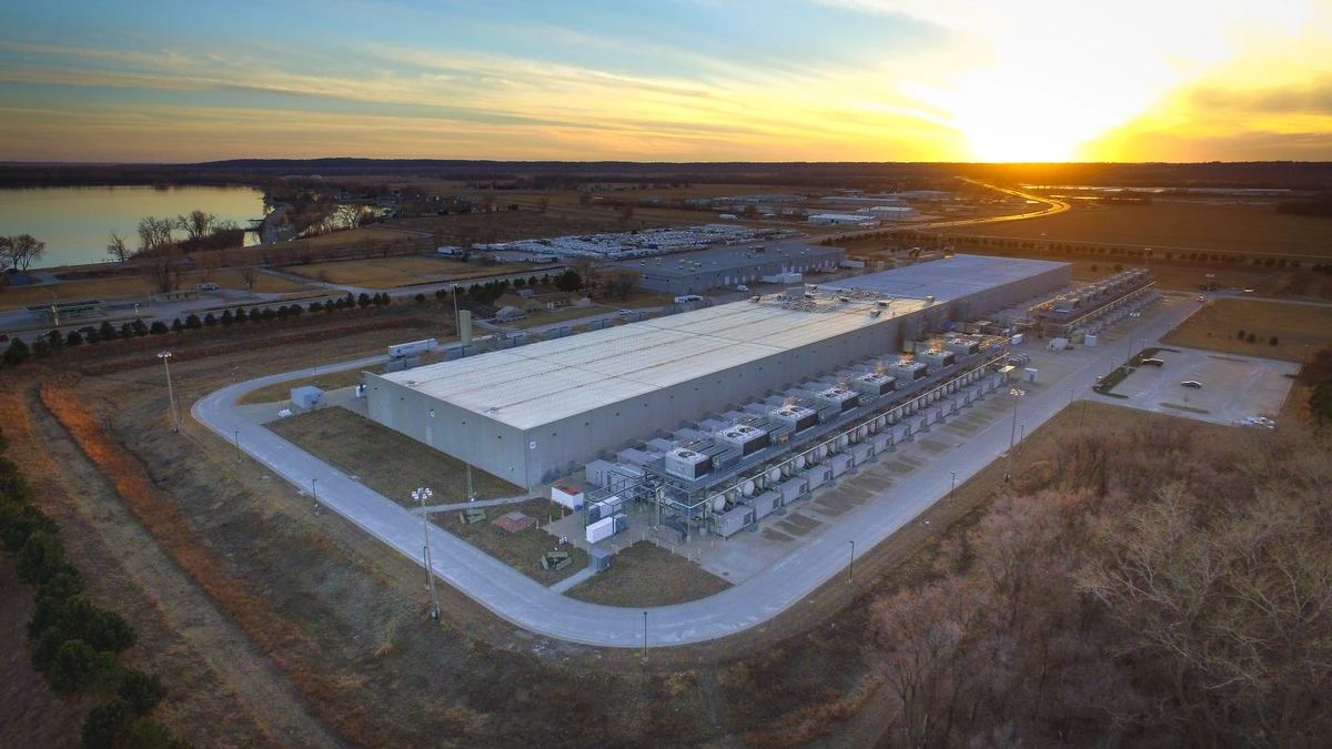 Aerial view at sunset of a long, flat grey Google data centre building with rows of cooling equipment along one side, surrounded by roads, bare trees and farmland beside a river