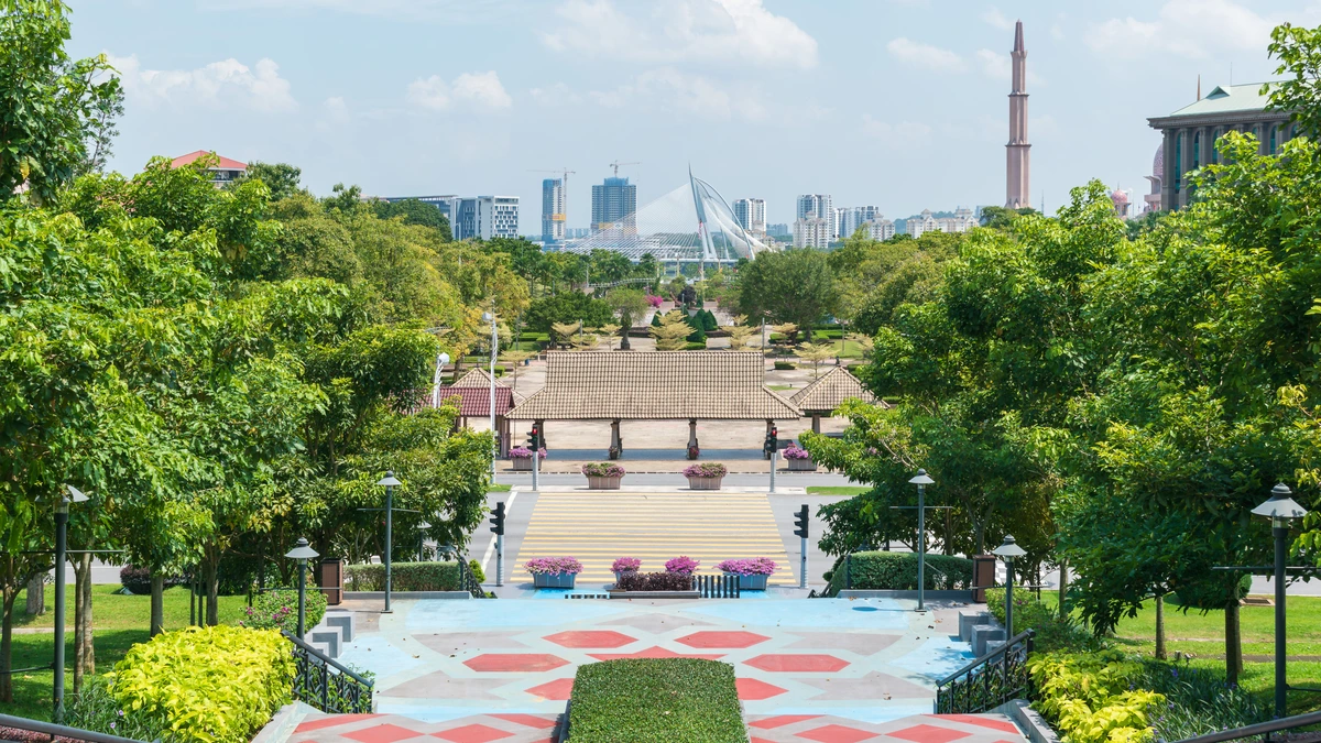 View from the Putrajaya Steps down a landscaped boulevard of trees and red and blue patterned paving towards the city centre skyline, with a cable-stayed bridge and a tall minaret in the distance