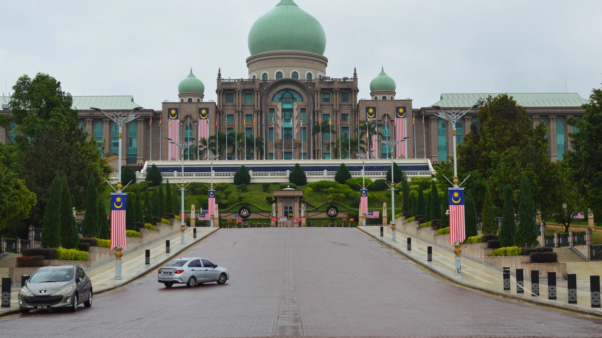 Perdana Putra in Putrajaya, a large building with a green central dome and smaller domes, at the end of a long approach road lined with Malaysian flags under an overcast sky
