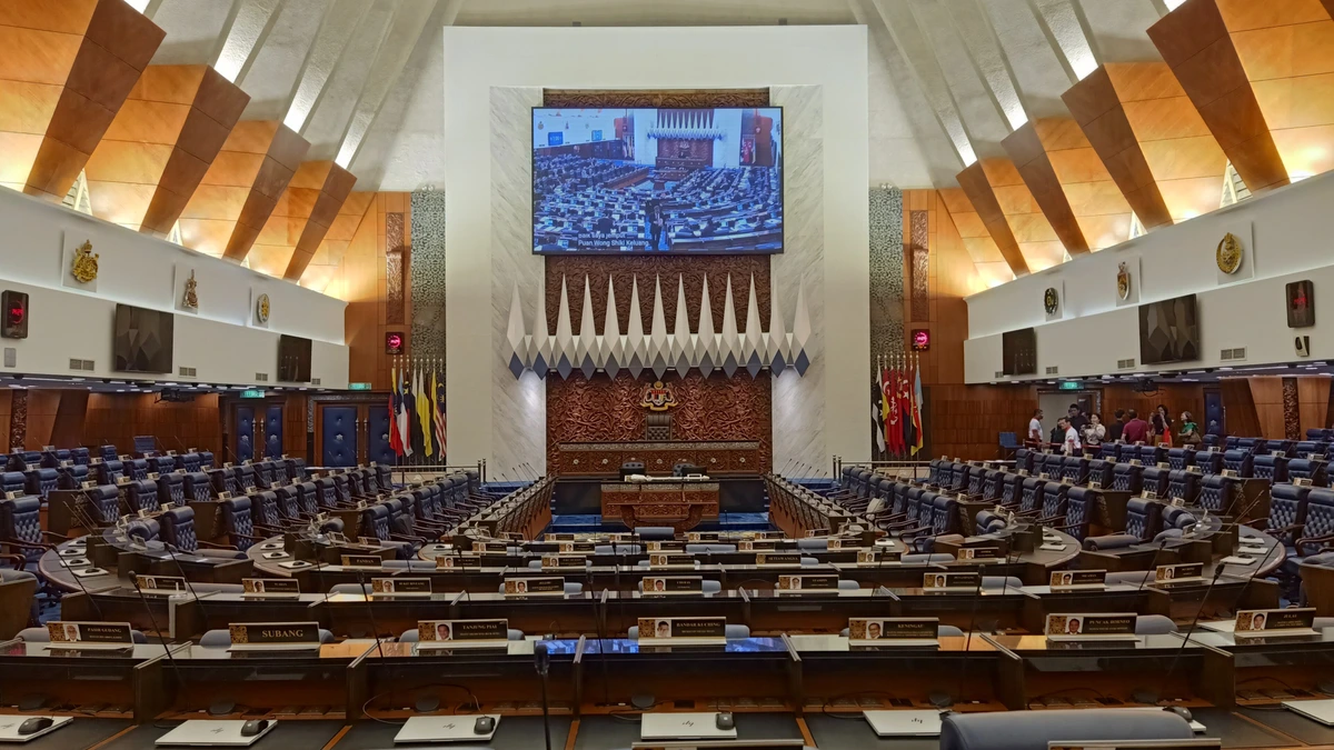 Front of the Dewan Rakyat chamber with the Speaker's chair, state crest, flags and a large screen, seen from the members' desks