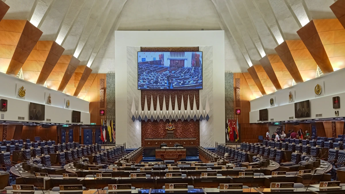 The Dewan Rakyat chamber in Malaysia's Parliament with rows of desks, name plates, flags and a large screen under an angular timber ceiling