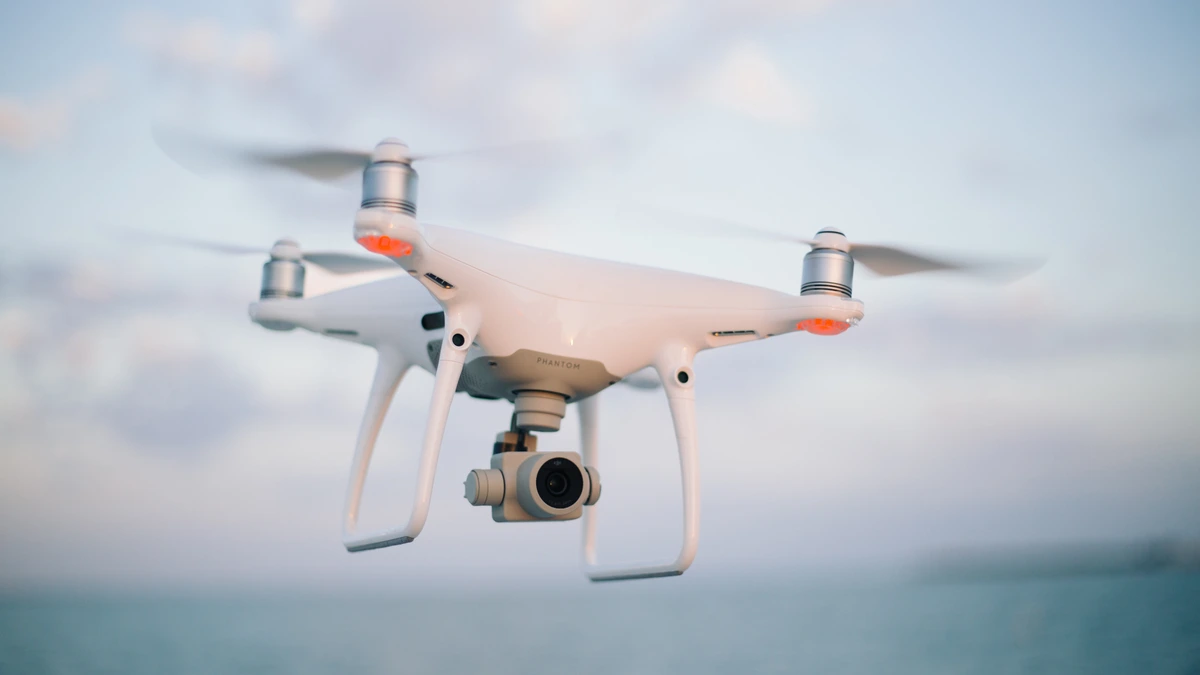 A white quadcopter drone with a gimbal camera hovering in flight over the sea under a soft sky
