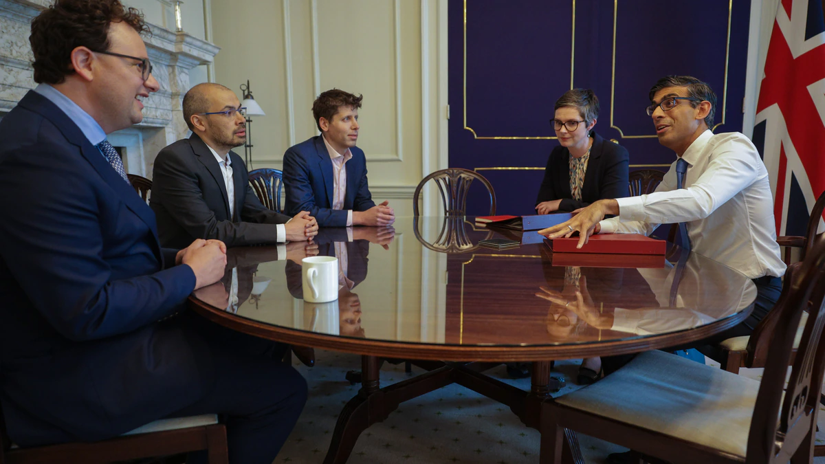 Five people in business dress sit around a polished oval table in a panelled state room, three on one side and two on the other, with a Union flag and a red ministerial box on the table