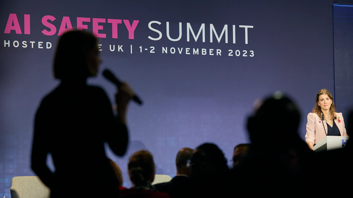 A government minister speaks from a lectern on a darkened stage in front of a large backdrop reading AI Safety Summit, hosted by the UK, 1 to 2 November 2023, with the silhouettes of a standing questioner and a seated audience in the foreground