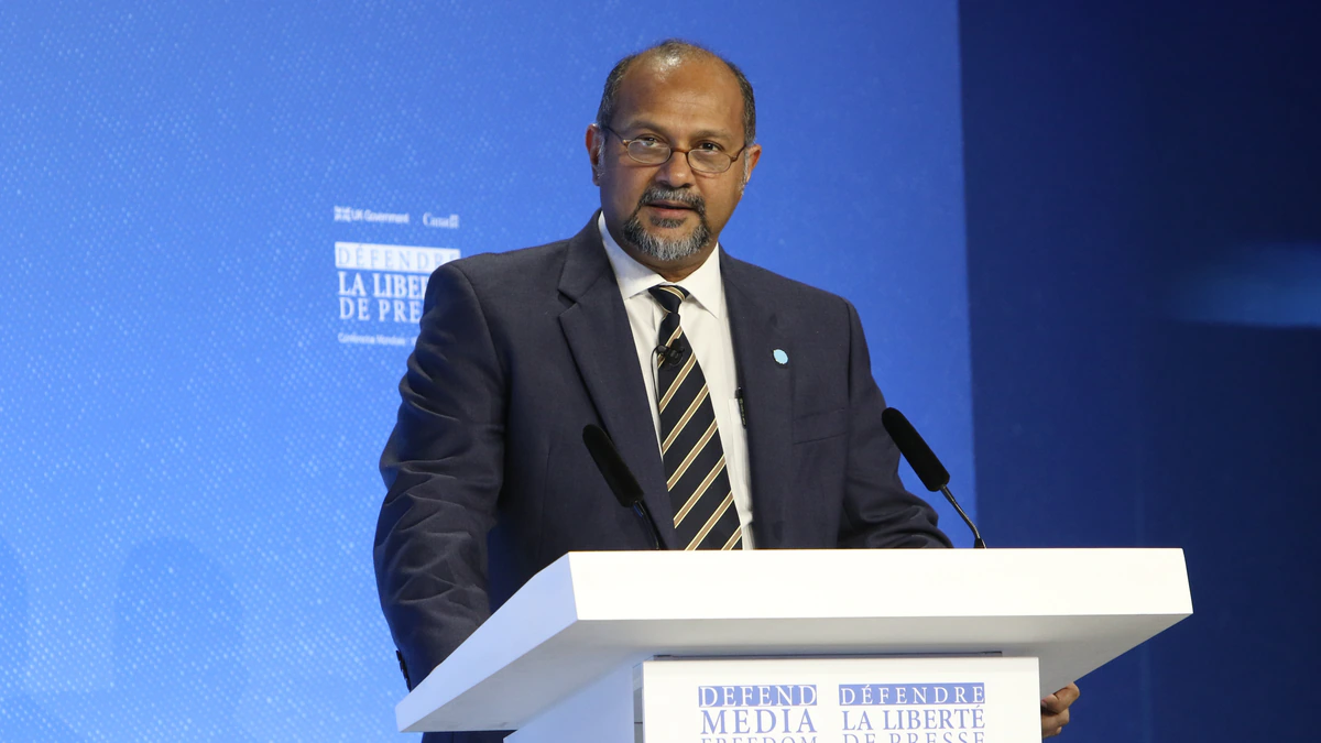 A bearded man in glasses and a dark suit stands at a white lectern marked Defend Media Freedom, speaking against a deep blue conference backdrop