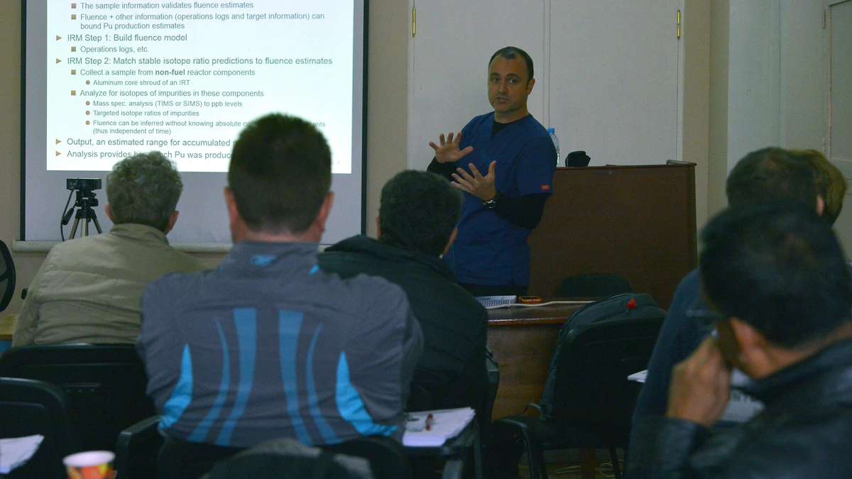 A trainer in blue scrubs gestures in front of a projected slide headed EDM Tool: Verification Mission while roughly eight seated trainees take notes in a plain classroom