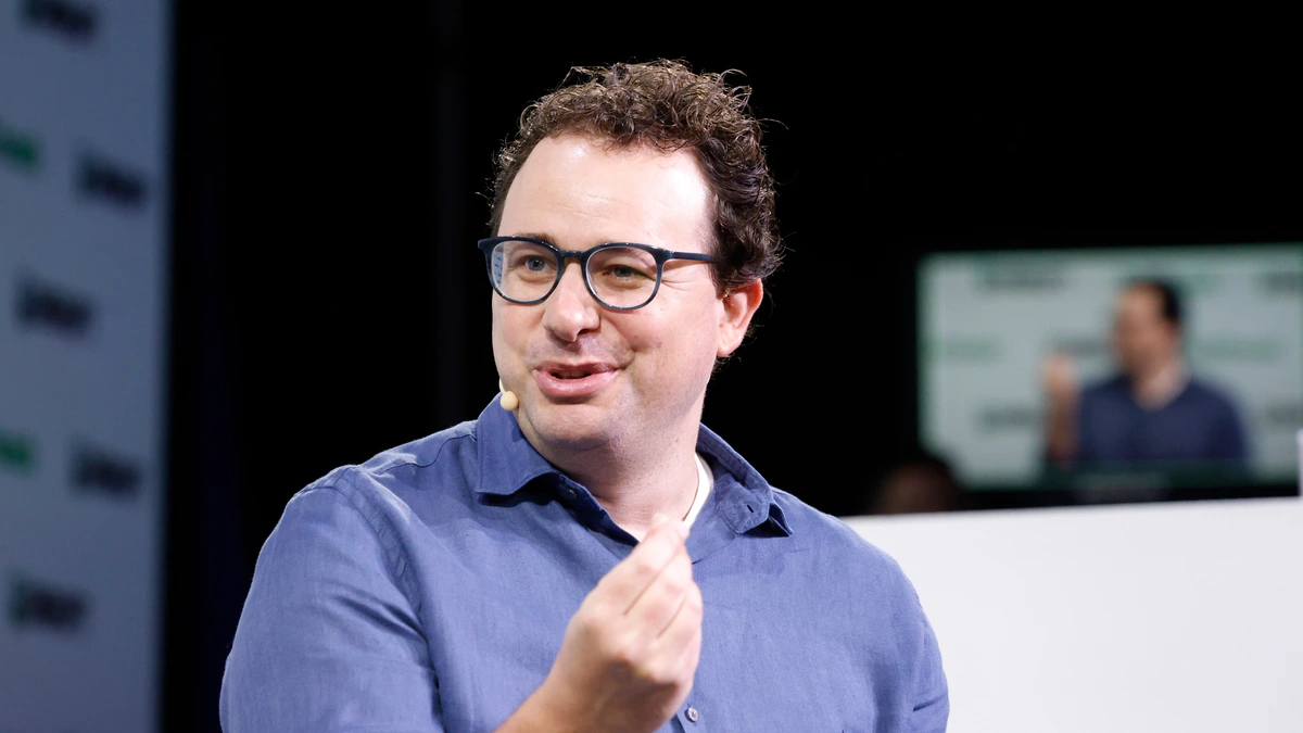 Dario Amodei, wearing glasses and a blue shirt, speaks on a dark stage while gesturing with one hand, with a blurred screen behind him
