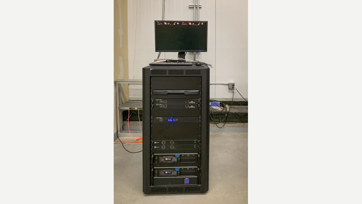 A single black server rack of stacked equipment with a small monitor on top, standing on a bare concrete floor against a white wall