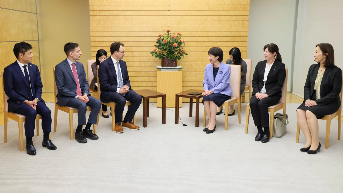 Two rows of chairs in a formal reception room with a pale wooden wall and a flower arrangement, with a group of men in suits seated facing Sanae Takaichi and officials