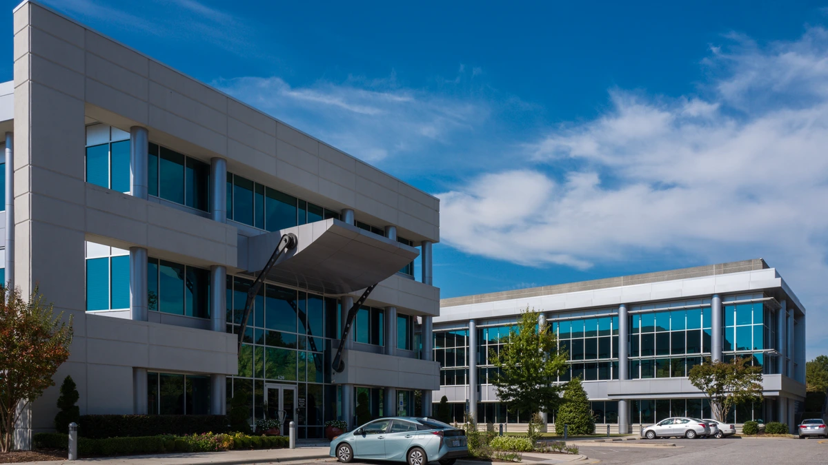 A three-storey office building with grey panels, tall glass windows and a cantilevered entrance canopy, with a car parked in front under a blue sky