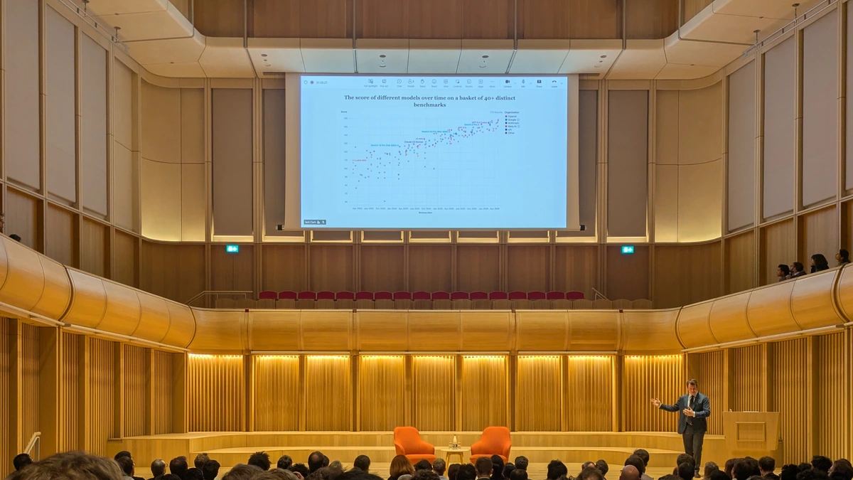 A speaker gestures on a wooden concert-hall stage beside two orange armchairs while a large screen above him shows a scatter chart of AI benchmark scores rising over time, with an audience in the foreground