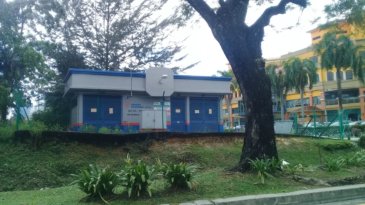 A small blue and grey Tenaga Nasional electrical substation building on a grassy bank behind a large tree