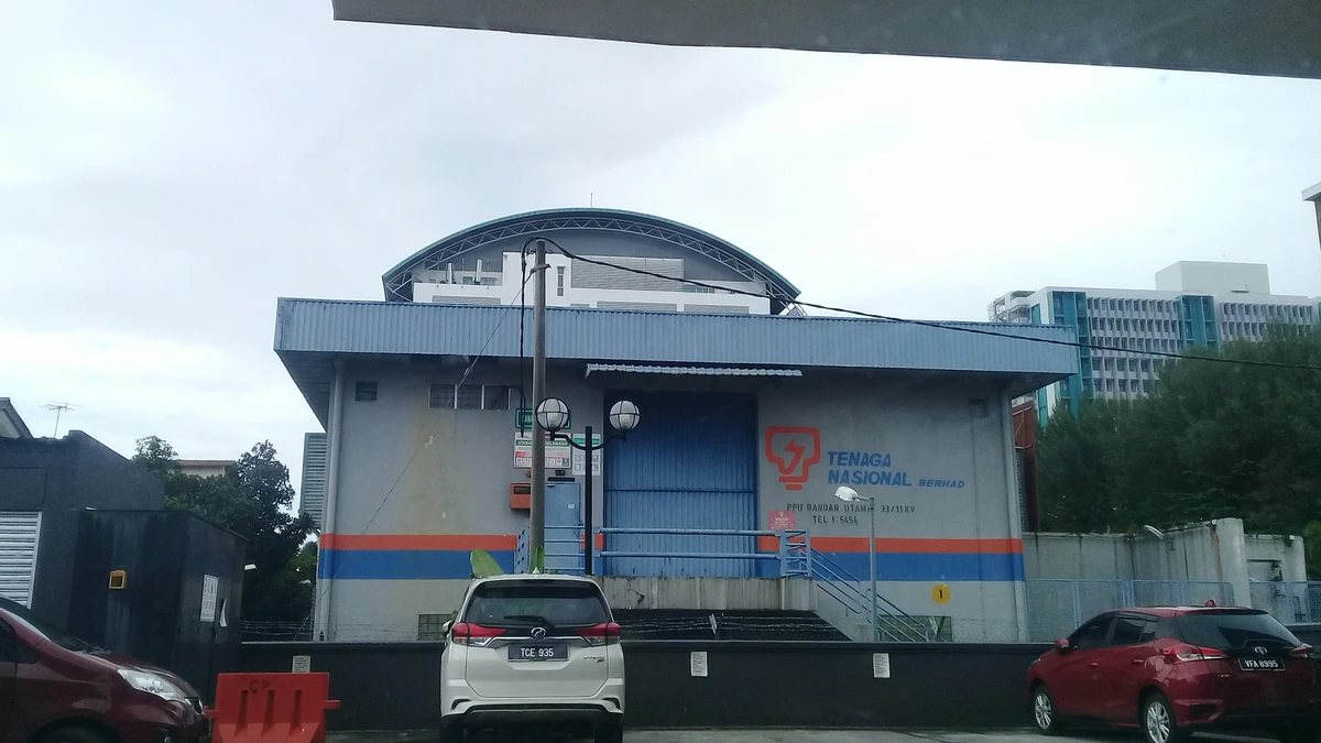 A grey and blue Tenaga Nasional main intake building with a curved roof and orange logo, with cars parked in front