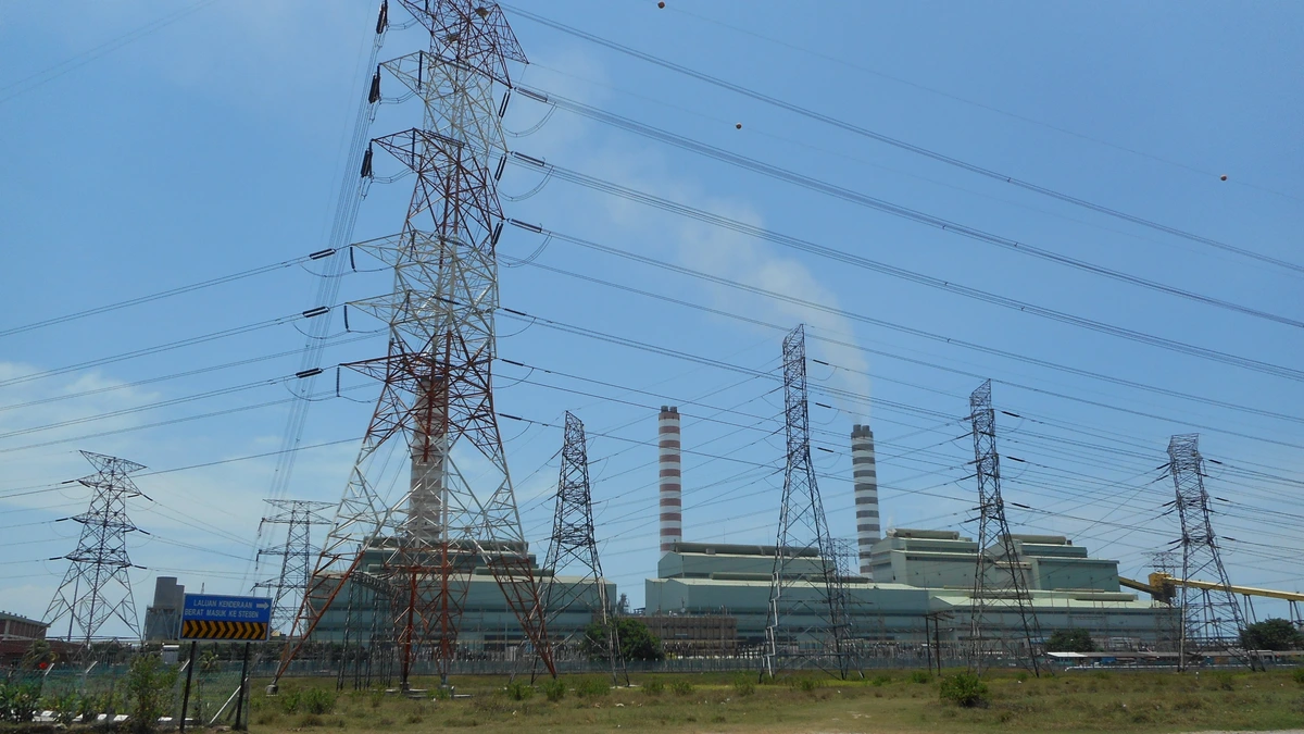 Steel transmission towers and power lines in front of a large coal-fired power station with two striped chimneys under a blue sky