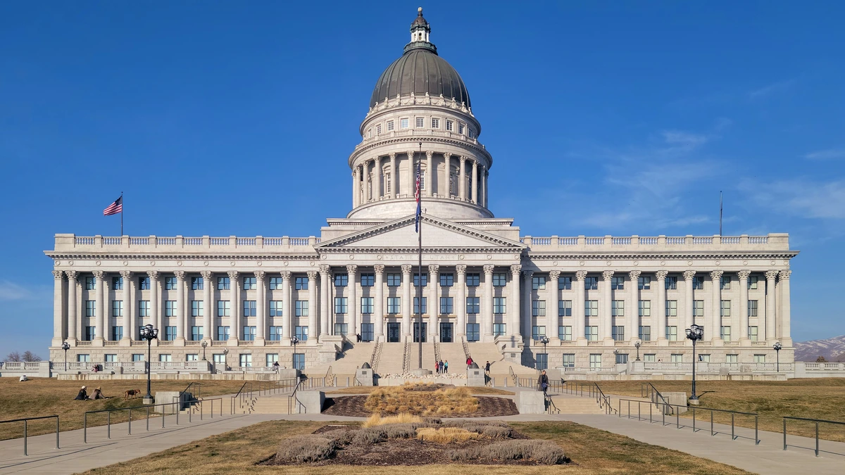 Utah State Capitol with a grey dome and white columns under a clear blue sky, seen from the south across a lawn and paved plaza