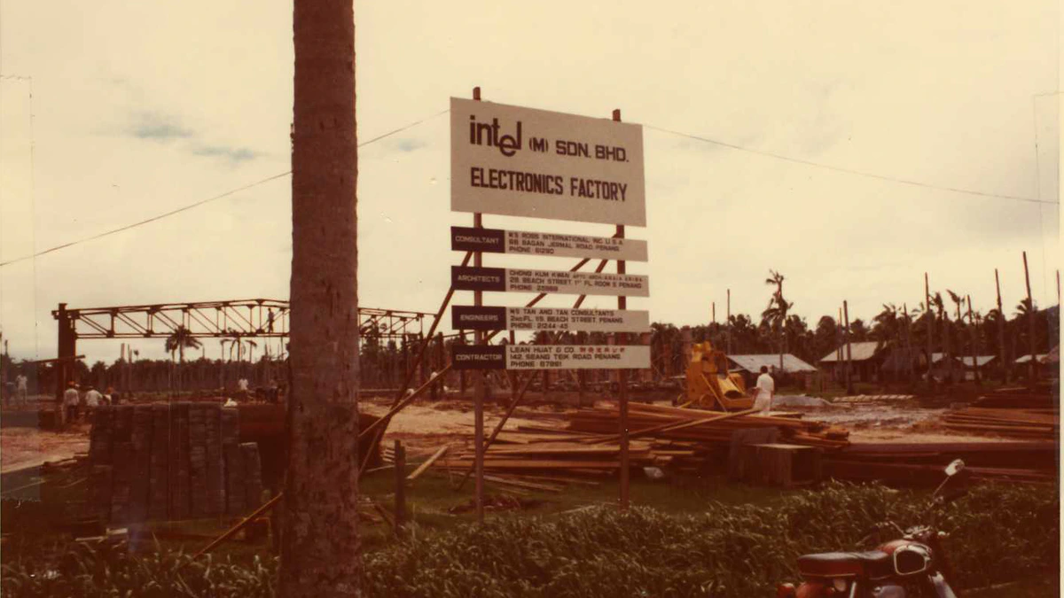 A faded colour photograph of a construction site with a large sign reading Intel (M) Sdn Bhd Electronics Factory, stacked timber, palm trees and a parked motorcycle