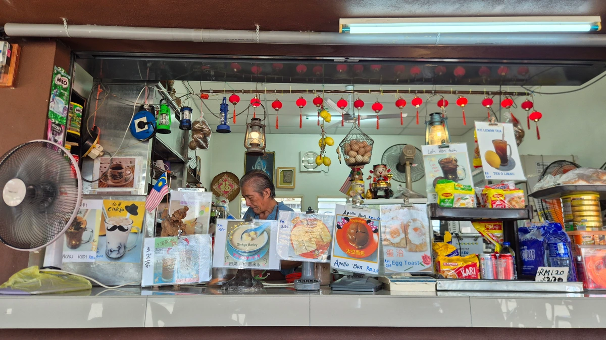 An older man works behind the counter of a small coffee stall in Batu Gajah, Perak, surrounded by hand-made menu cards for kopi, Apollo buns and egg toast, with red lanterns hanging above and a small Malaysian flag at left