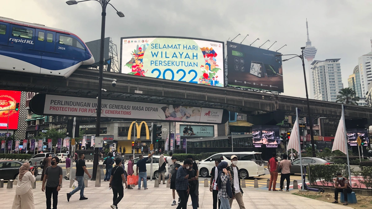 Pedestrians cross the plaza at the Jalan Bukit Bintang and Jalan Sultan Ismail junction in Kuala Lumpur beneath large billboards, including a Federal Territory Day hoarding and a laptop advertisement, with a monorail train on the elevated track at left