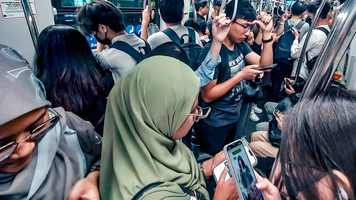 Commuters crowd a Kuala Lumpur rail carriage during the evening rush, several looking down at their smartphones, with a woman in a green headscarf in the foreground and a lit phone screen at lower right
