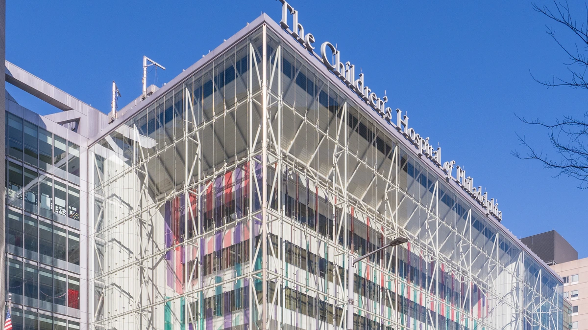 A tall glass-fronted hospital building with a white steel lattice and coloured panels behind the glass, its roofline sign reading The Children's Hospital of Philadelphia, under a clear blue sky