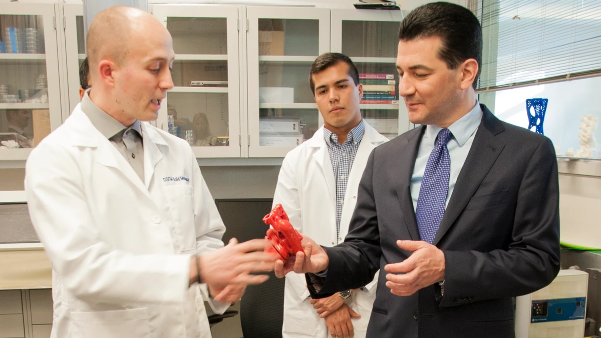 A man in a white lab coat hands a small red 3D-printed heart model to a man in a dark suit and blue tie, while a younger man in a lab coat watches in a laboratory