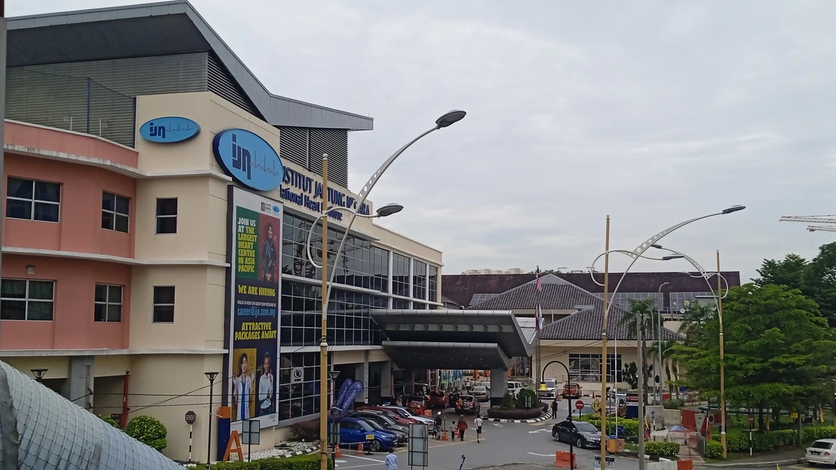 The pink and cream Institut Jantung Negara building with blue IJN logos and a hiring banner, seen from a footbridge above rows of parked motorcycles on Jalan Tun Razak