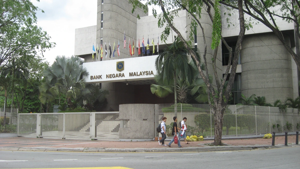 The concrete frontage of the Bank Negara Malaysia headquarters in Kuala Lumpur behind a row of flagpoles, with a long sign reading BANK NEGARA MALAYSIA above the entrance and pedestrians passing on the pavement