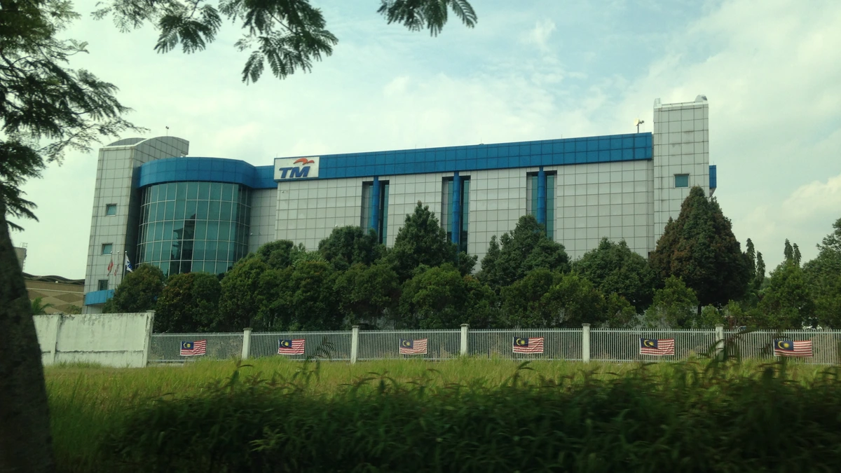 A low blue and grey Telekom Malaysia building behind a line of trees and a perimeter fence hung with Malaysian flags, with tall grass in the foreground