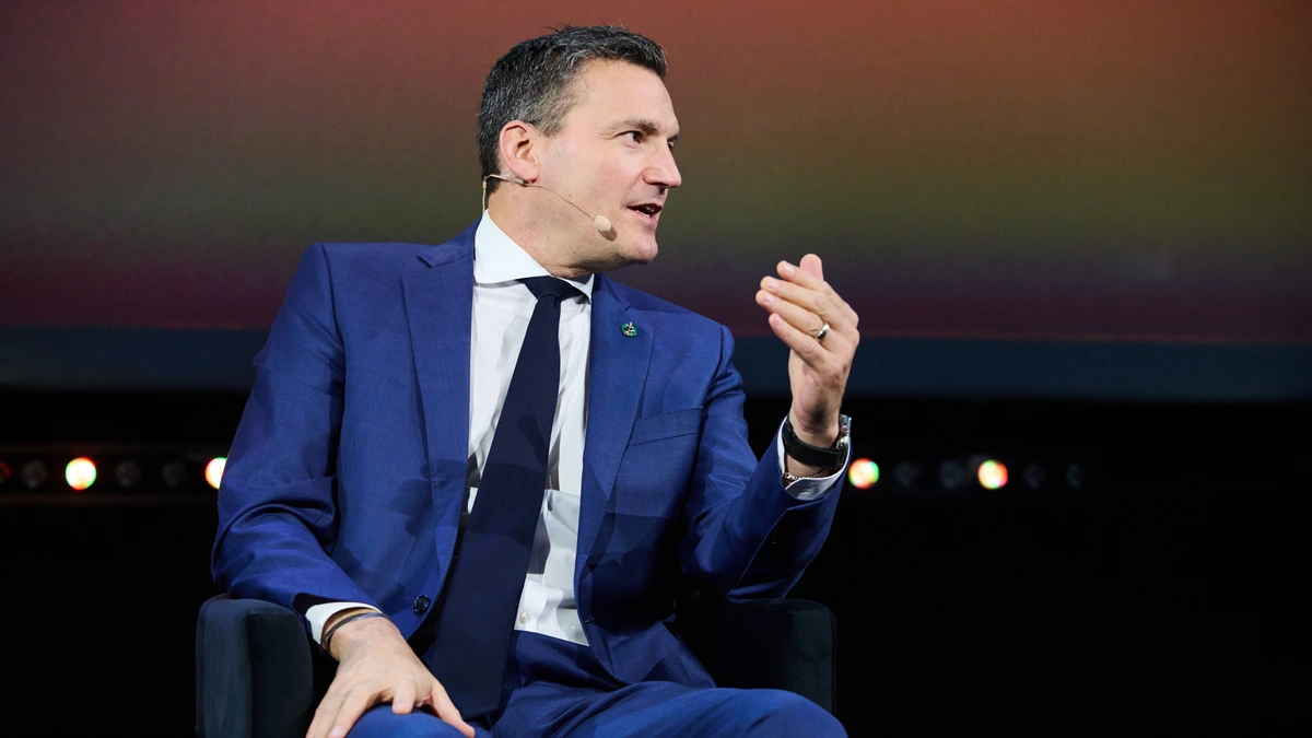 Evan Solomon in a dark blue suit and navy tie, seated on a low stage chair with one hand raised mid-sentence, against a warmly lit conference backdrop