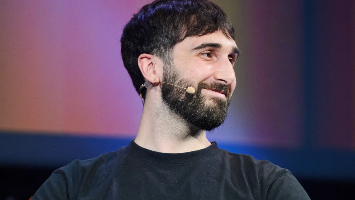 Cohere co-founder and chief executive Aidan Gomez in a black T-shirt and a thin headset microphone, smiling and looking off to one side in front of a softly lit purple and orange stage backdrop