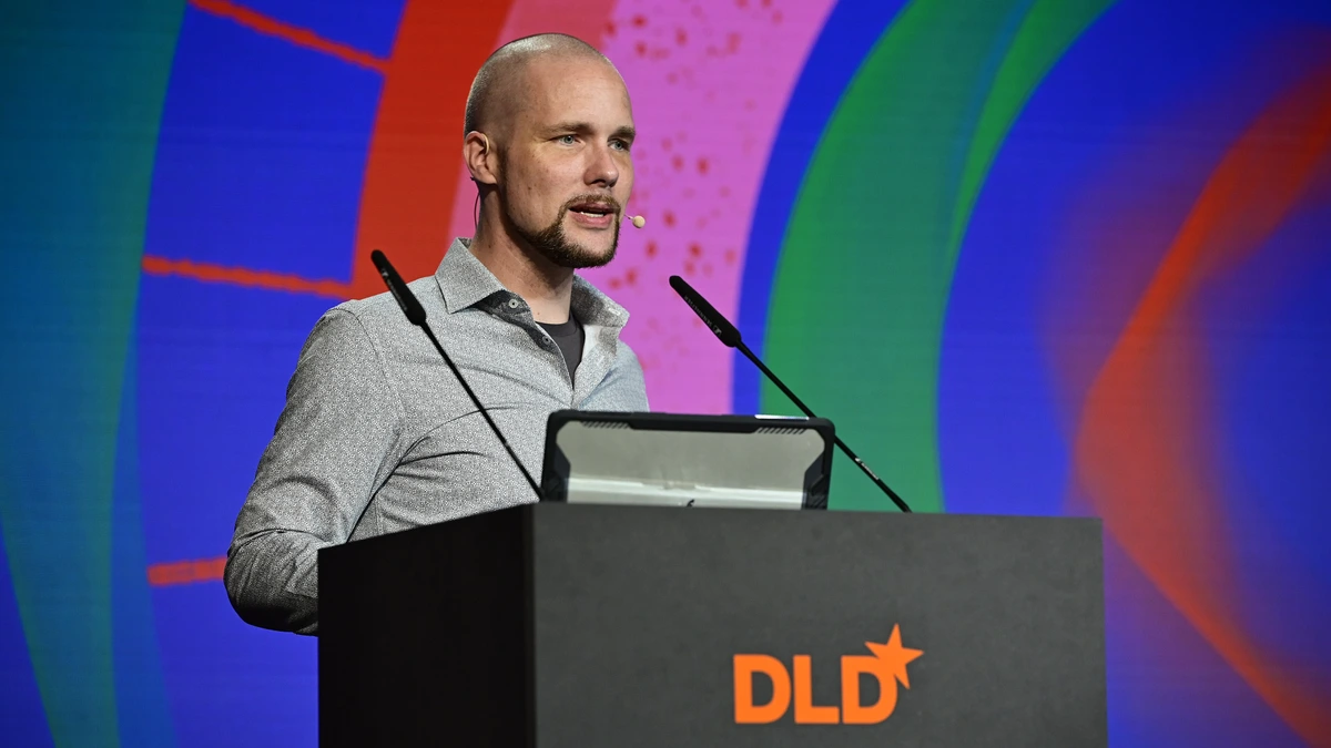 Jonas Andrulis in a grey shirt at a black lectern carrying the orange DLD logo, speaking into a headset microphone in front of a brightly coloured abstract screen