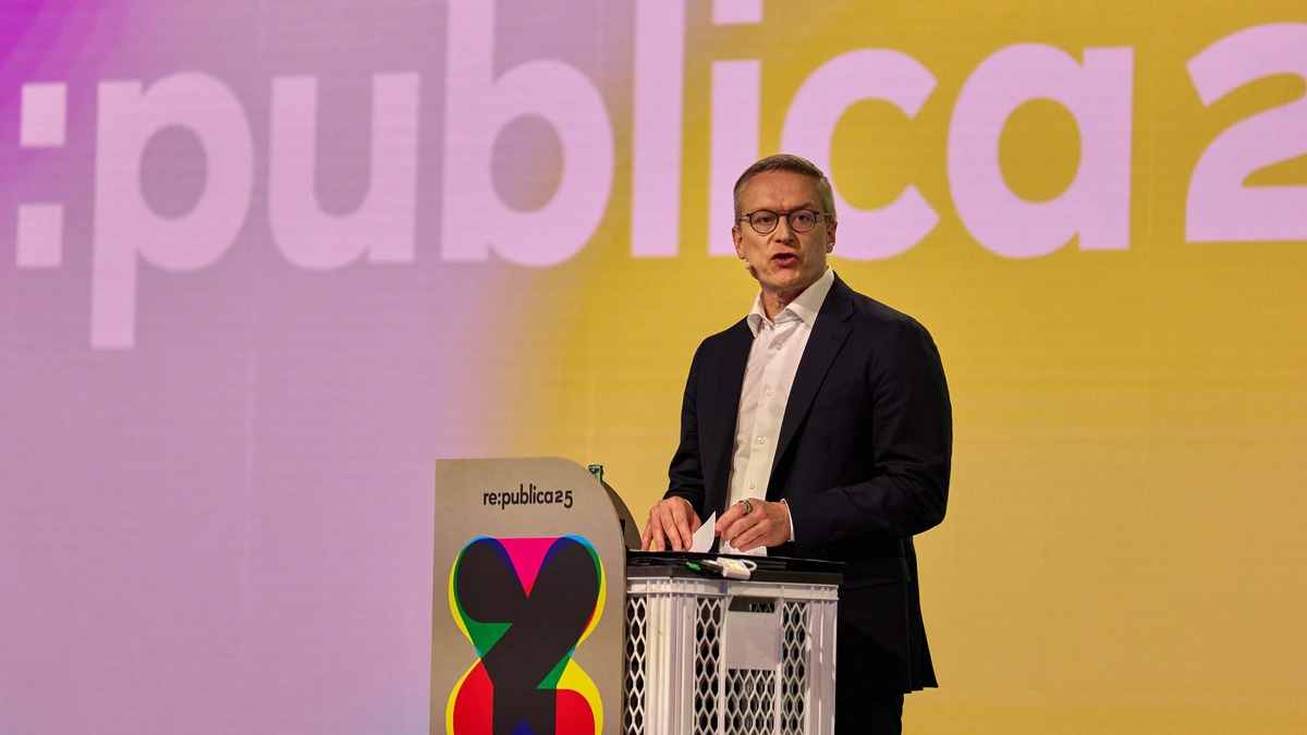 Karsten Wildberger standing at a white lectern marked re:publica 25, speaking in a dark jacket in front of a large pink and yellow conference screen