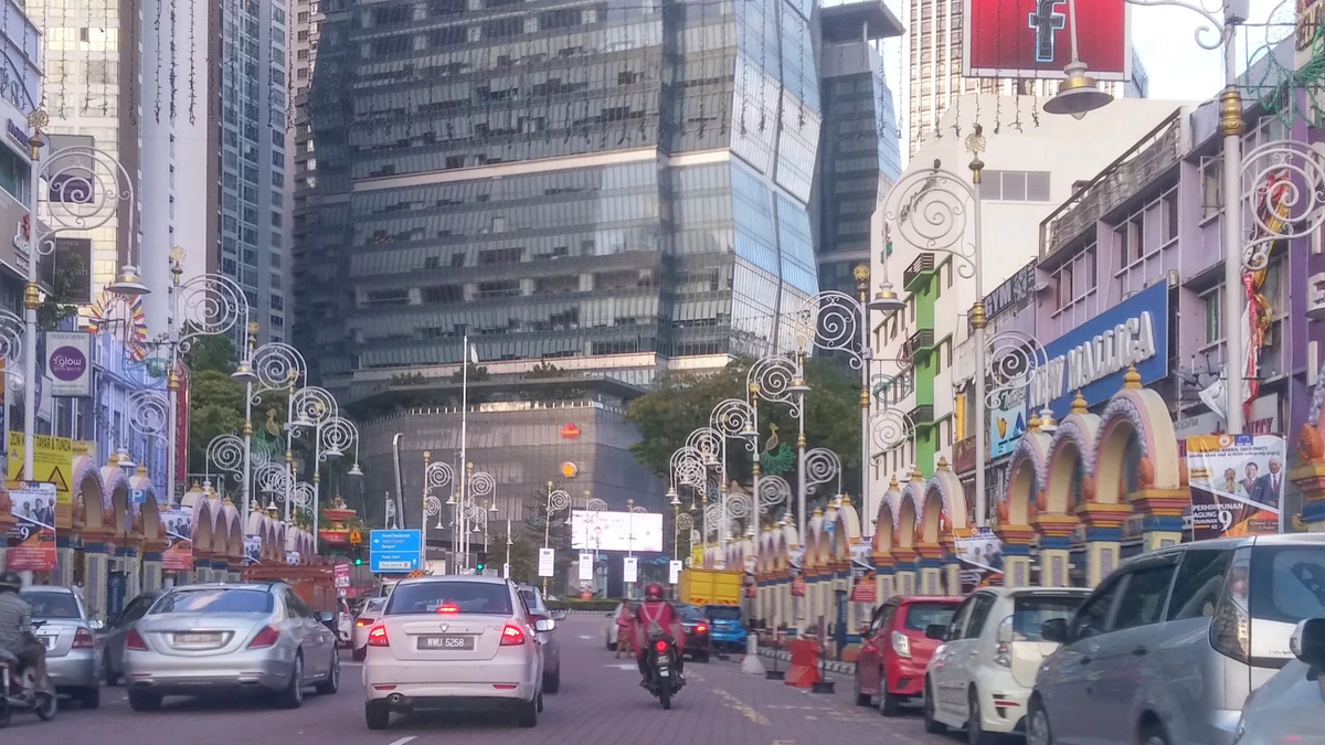 Traffic on a divided road through Brickfields in Kuala Lumpur, lined with decorative arches and ornamental street lamps, shopfronts on both sides and glass office towers rising behind
