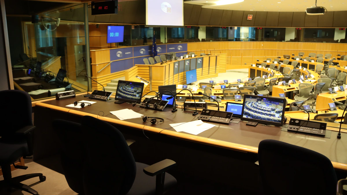 The view from inside an empty simultaneous interpreting booth, with headphones, papers and audio consoles on the desk and a glass wall looking out over a tiered European Parliament committee room
