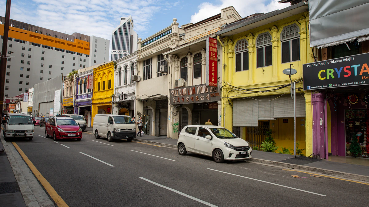 A row of restored shophouses on a Kuala Lumpur street, their upper floors painted yellow and cream, with vertical Chinese shop signs, a Malay-language signboard and parked vans along the kerb