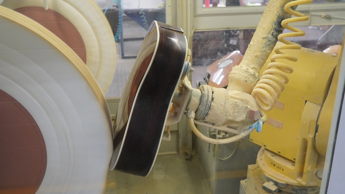 A yellow industrial robot arm holds a guitar body against a spinning polishing wheel inside a dusty booth