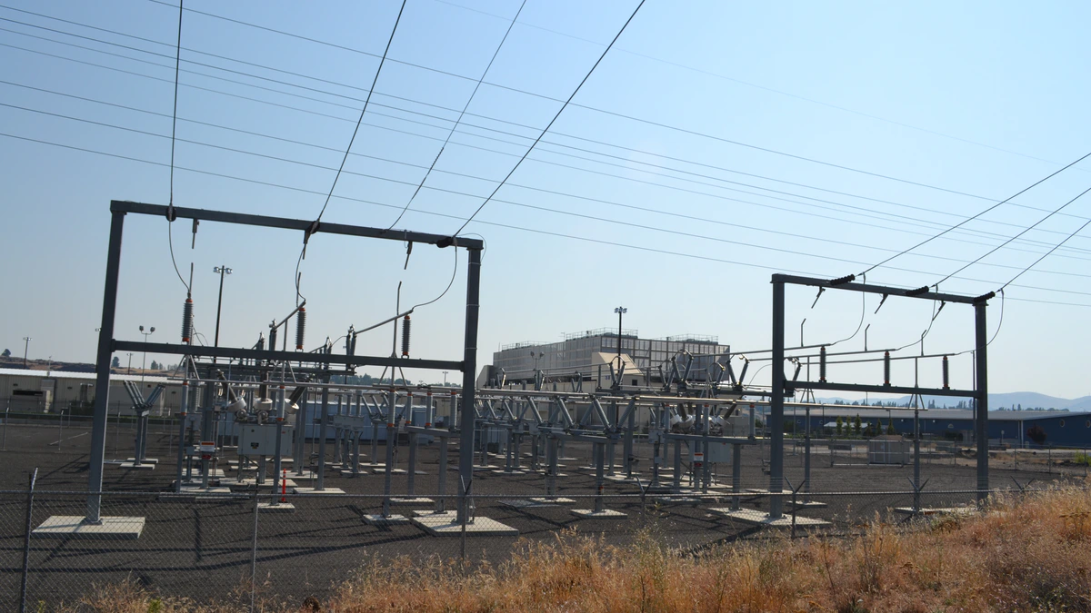 An electrical substation of steel gantries and insulators behind a chain-link fence, with the long low sheds of a Google data centre and its rooftop cooling plant visible beyond