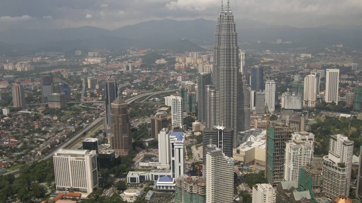 An aerial view across central Kuala Lumpur with the Petronas Twin Towers rising above office blocks, elevated highways and dense housing, hills fading into haze on the horizon