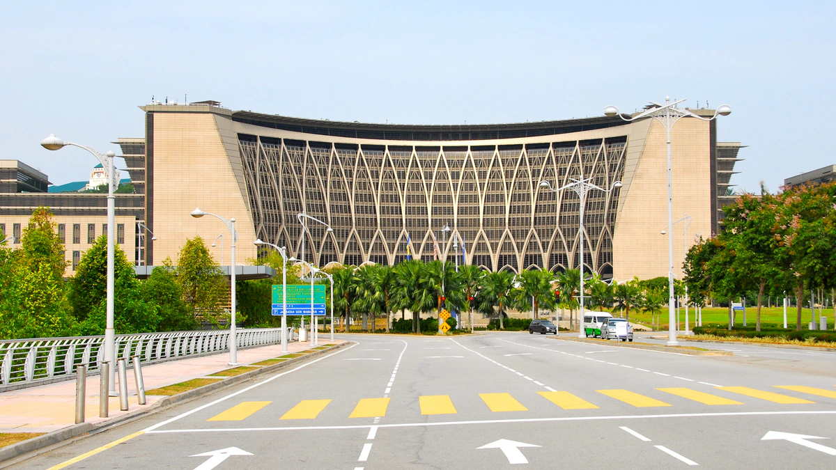 A wide curved federal government building in Putrajaya with a pale stone facade and a tall lattice of pointed arches across its front, seen from an empty multi-lane road lined with palms and street lamps