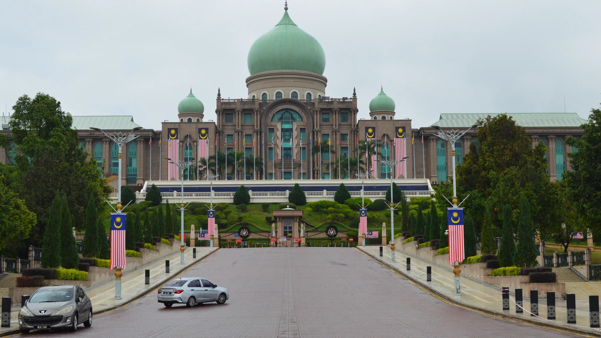 Green-domed government complex with Malaysian flags on lamp posts at the top of a wide, wet approach road